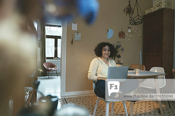 Smiling woman with laptop sitting cross-legged on chair at home