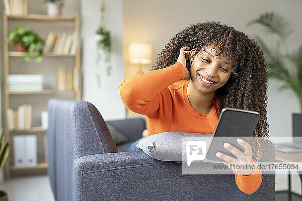 Happy young woman with hand in hair using mobile phone on sofa at home
