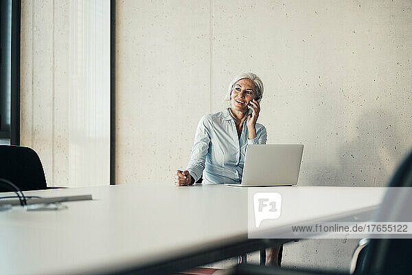 Lächelnde Geschäftsfrau  die mit einem Laptop am Schreibtisch sitzt und mit einem Smartphone spricht