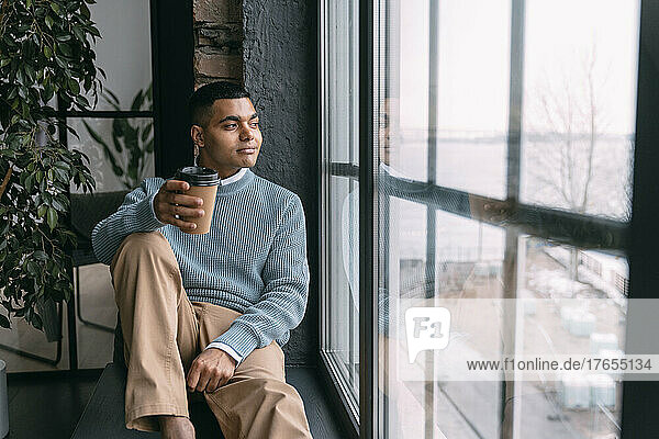 Young man with disposable coffee cup looking through window sitting at home