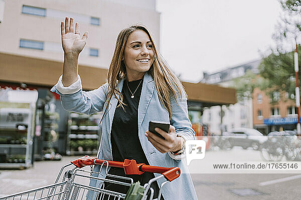 Smiling woman with smart phone waving at someone standing by store