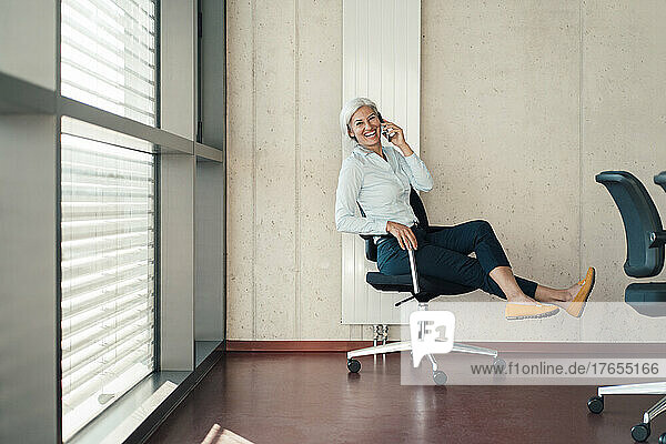 Cheerful businesswoman talking on mobile phone sitting on chair at work place