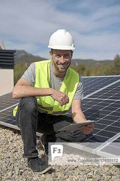 Smiling engineer with tablet PC crouching by solar panels on sunny day
