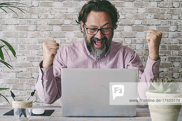 Happy businessman gesturing fist sitting with laptop at home