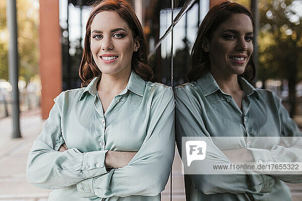 Smiling businesswoman with arms crossed leaning on glass wall