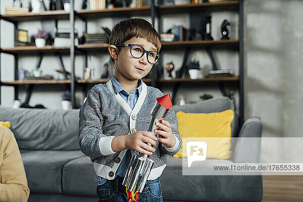 Cute boy wearing eyeglasses holding toy rocket at home