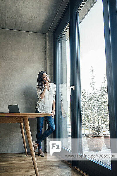 Smiling woman talking on mobile phone looking through window at home