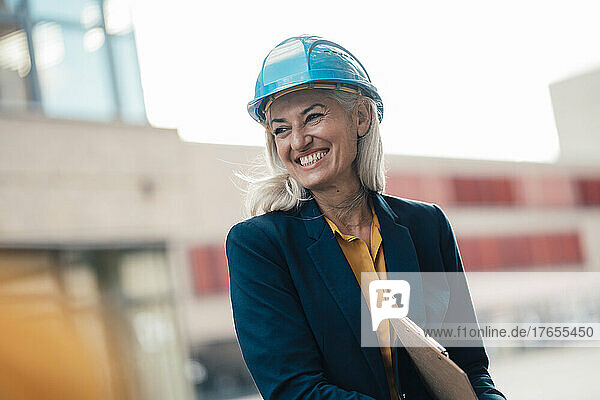 Cheerful female architect with clipboard wearing hardhat at office park