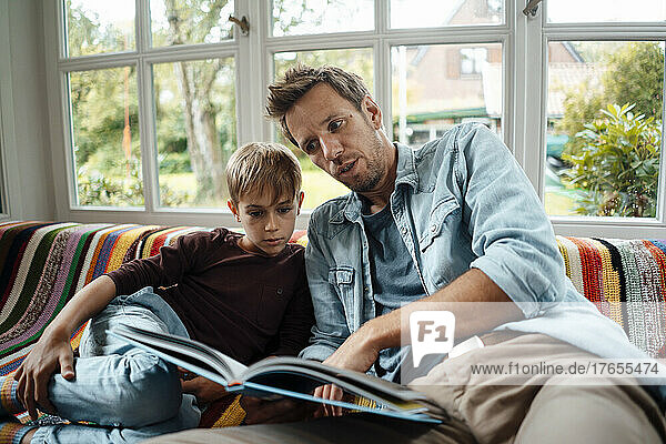 Man reading book to son sitting on sofa at home