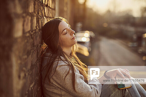 Young woman leaning on brick wall with eyes closed