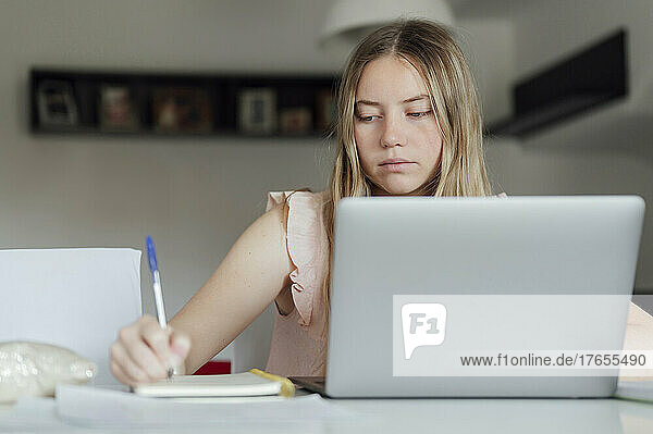 Young woman writing on note pad sitting with laptop at home