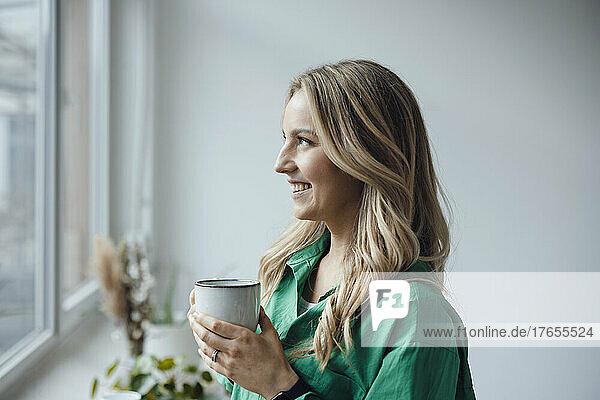 Lächelnde Frau mit Kaffeetasse und Blick aus dem Fenster zu Hause