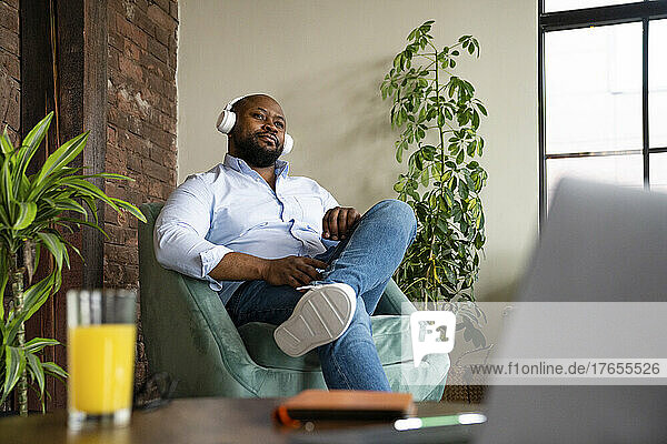 Businessman listening music through wireless headphones sitting on armchair in office