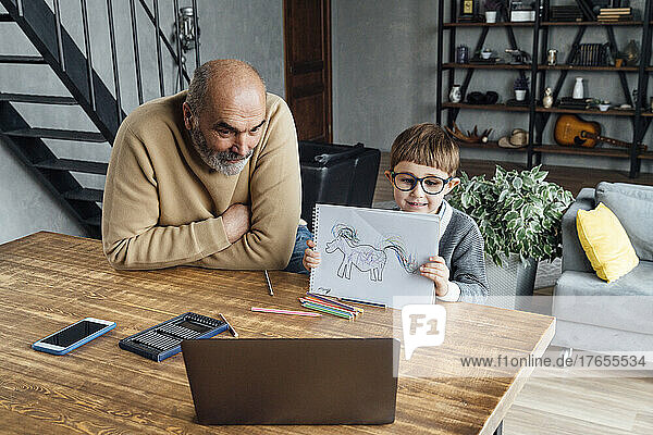 Cute boy showing drawing through laptop sitting by grandfather at home