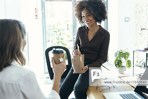 Smiling young businesswoman with take out food looking at colleague holding disposable cup in office