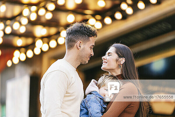 Smiling young woman carrying son looking at man in front of illuminated ceiling