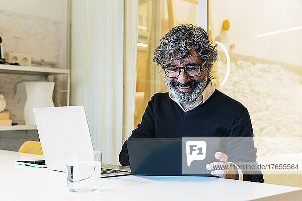 Happy senior freelancer using tablet PC sitting with laptop at table