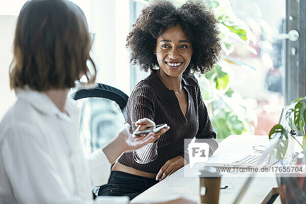 Lächelnde Geschäftsfrau gibt einem Kollegen am Schreibtisch im Büro ein Mobiltelefon