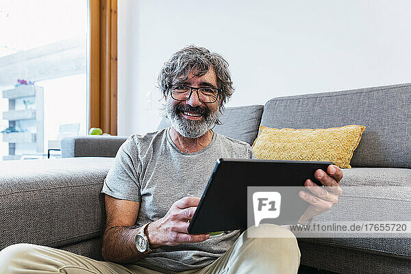 Smiling senior man with tablet PC sitting by sofa at home