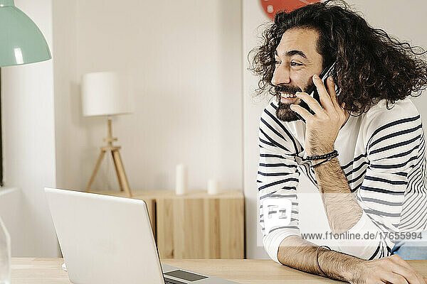Cheerful man talking on phone with his laptop next to him at home