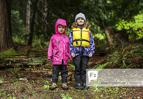 Two young girls in rain gear stand in a forest
