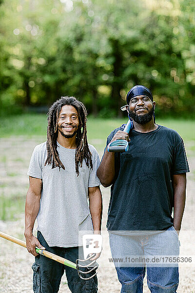 Two black farmers smiling at the camera while holding farm tools