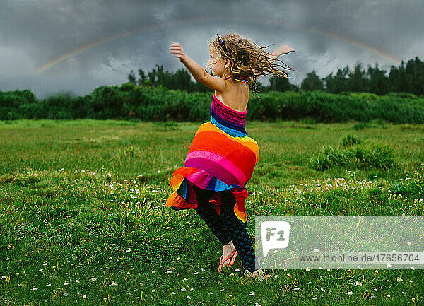 Child blond girl twirls in rainbow dress under a rainbow