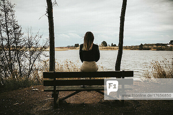 unrecognizable sad girl sitting on a bench while looking at a lake