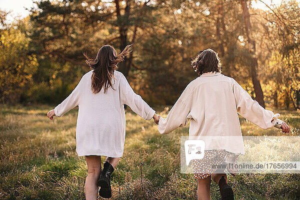 Back view of girlfriends holding hands while running in forest