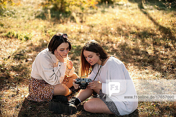 Girlfriends with camera sitting on grass in park in autumn