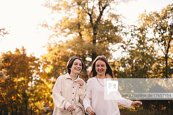 Two happy female friends smiling while walking in park during autumn