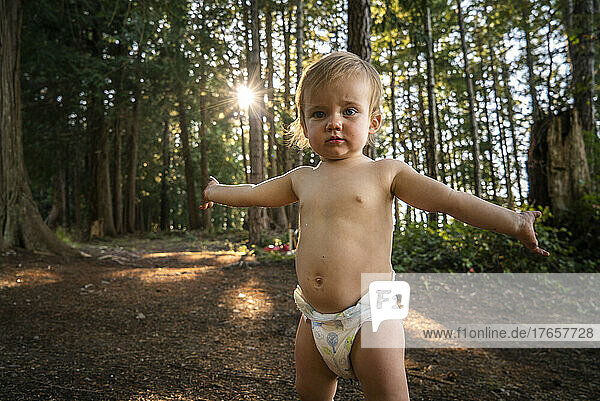 Baby in diaper stands with arms outstretched in forest
