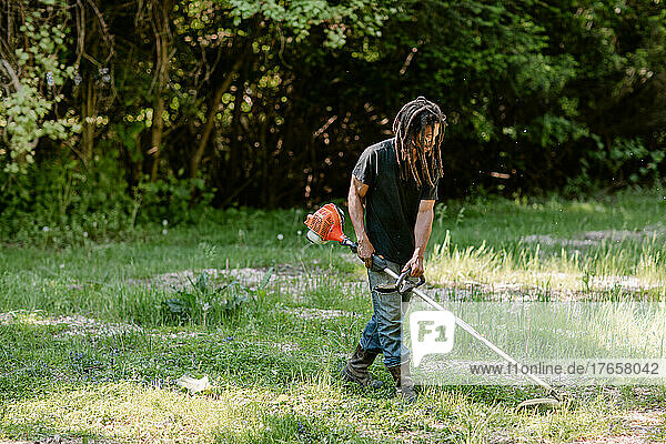 Black man with dreadlocks using a weed wacker on his farm