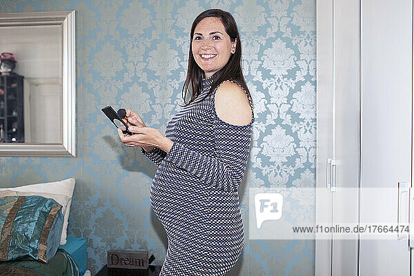 Porträt einer lächelnden schwangeren Frau  die sich im Schlafzimmer schminkt