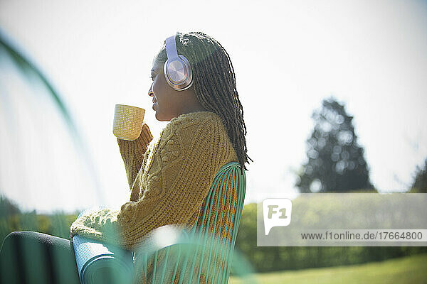 Junge Frau mit Kopfhörern und Yogamatte trinkt draußen Kaffee