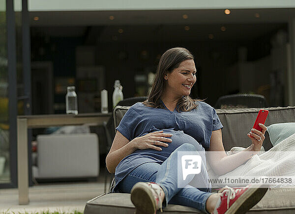 Glückliche schwangere Frau mit Smartphone auf einem Liegestuhl auf der Terrasse