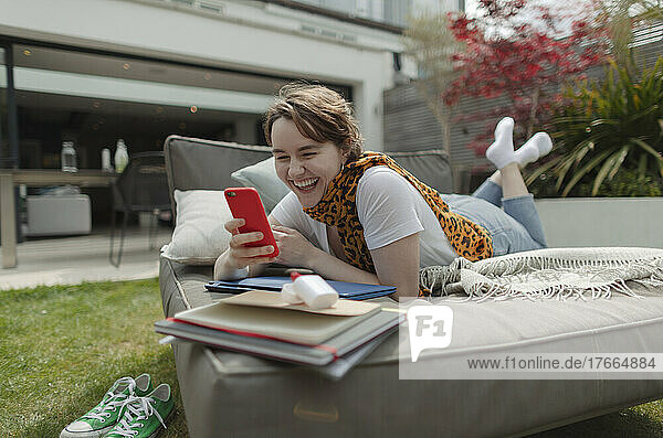 Glückliche junge Frau  die ein Smartphone auf einem Liegestuhl auf der Terrasse benutzt