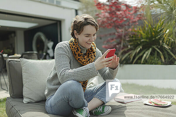 Lächelnde junge Frau  die ihr Smartphone auf einem Liegestuhl auf der Terrasse benutzt