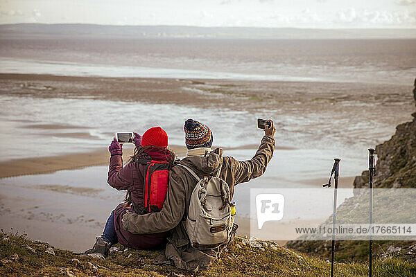 Hiker couple with camera phones on cliff overlooking winter beach