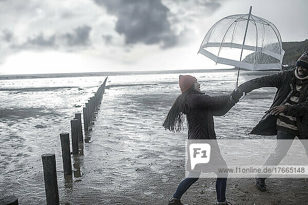 Verspieltes Paar mit Regenschirm am nassen Winterstrand