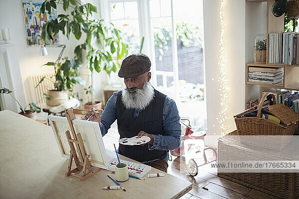 Älterer Mann mit Bart und Palette malt an der Staffelei