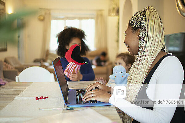 Mother working at laptop and watching daughters playing with puppets