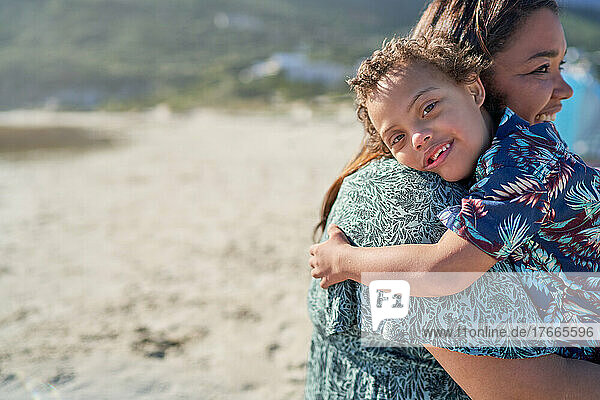Portrait happy boy with Down Syndrome over shoulder of mother on beach