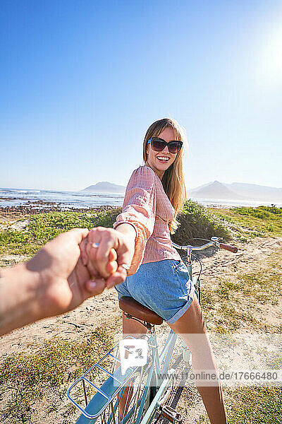 POV happy woman riding bicycle holding hand on sunny beach path