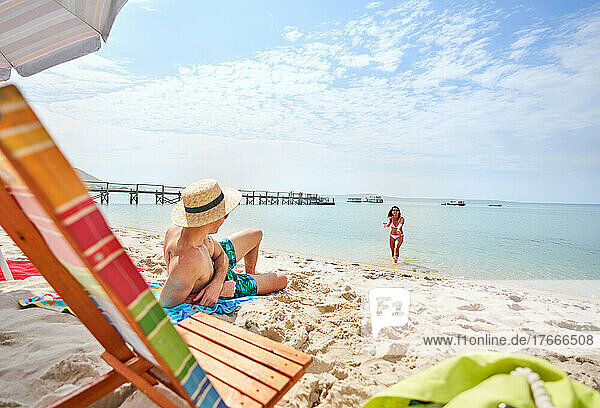 Girlfriend gesturing to boyfriend relaxing on sunny summer ocean beach