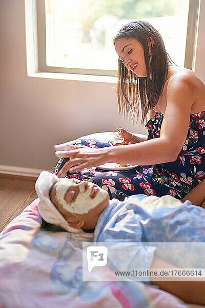 Mother giving daughter laying on bed a facial