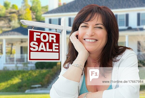 Frau mittleren Alters vor einem Haus mit einem Immobilienschild zum Verkauf im Hof