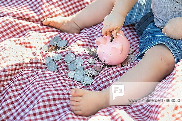 Baby boy sitting on picnic blanket putting coins in piggy bank