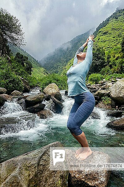 Junge  sportliche  fitte Frau macht Yoga Asana Utkatasana (Stuhlhaltung) im Freien an einem tropischen Wasserfall auf einem Stein stehend