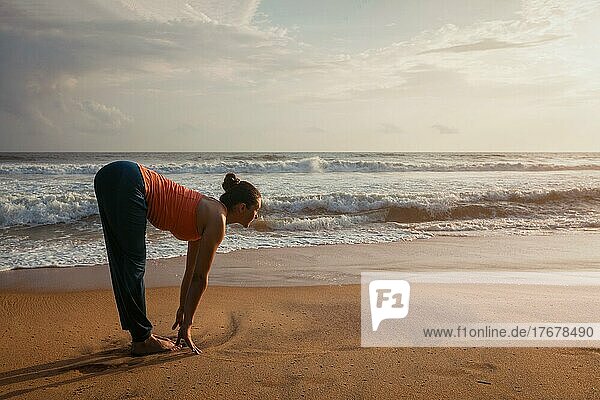 Junge sportliche fit Frau tun Yoga Sonnengruß Surya Namaskar Pose Uttanasana auf tropischen Strand bei Sonnenuntergang. Mit Linsenreflexion